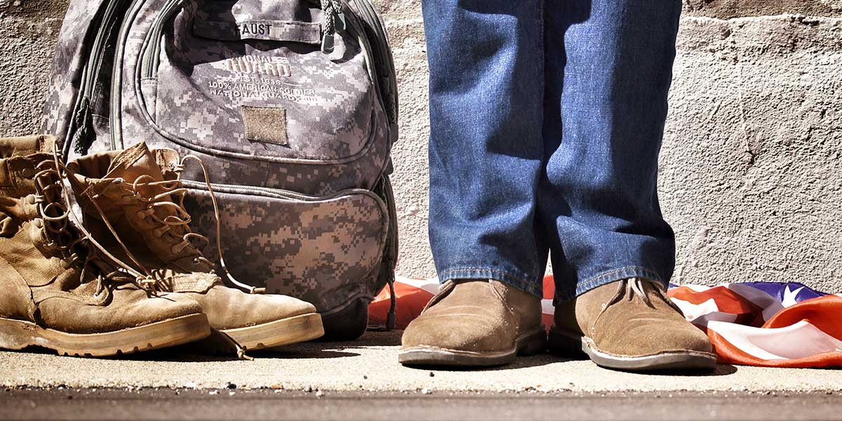 Veteran standing with a camouflage backpack, boots, and an American flag close to them