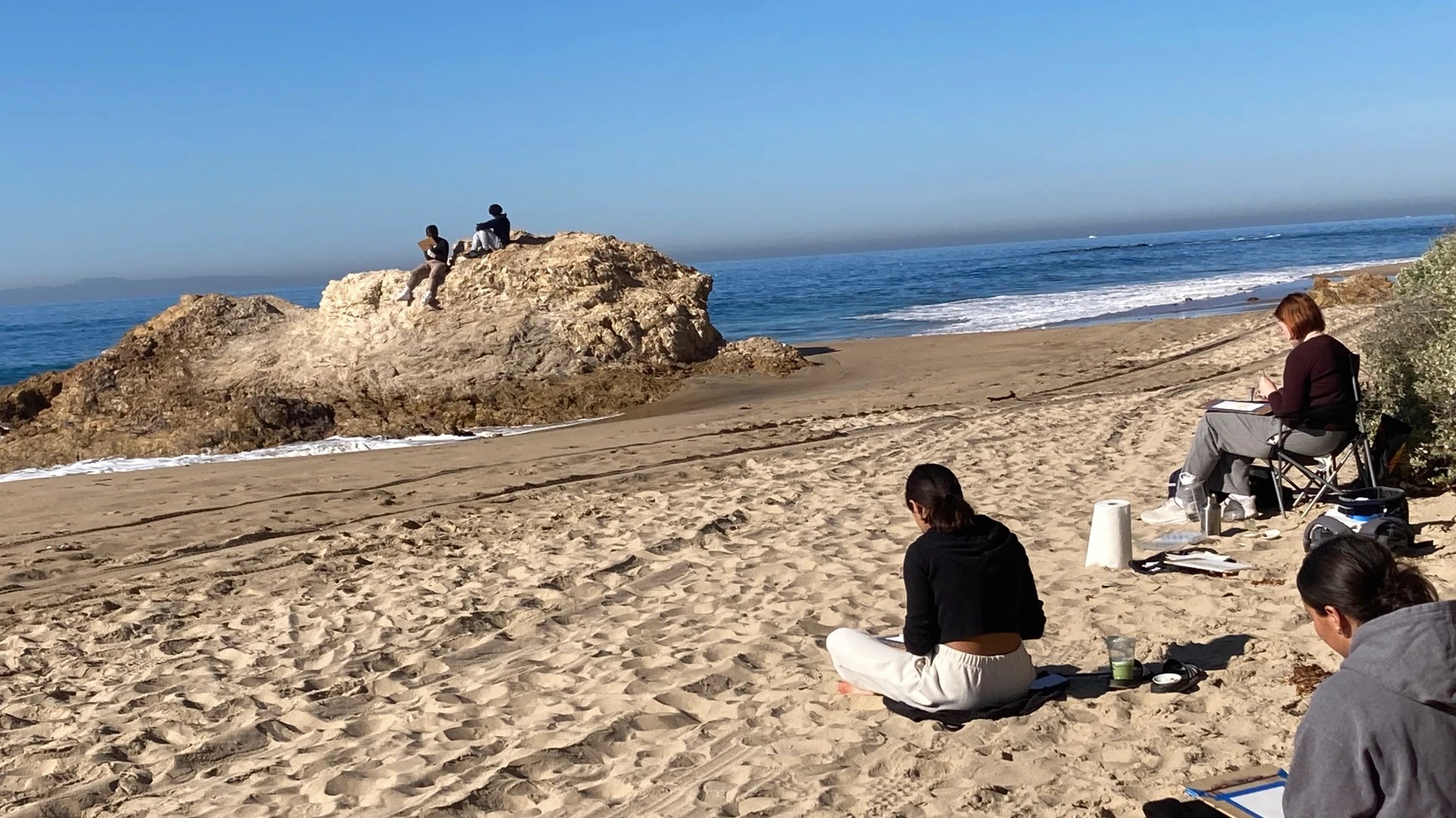 A woman sitting on a beach with a large rock and people sitting on it