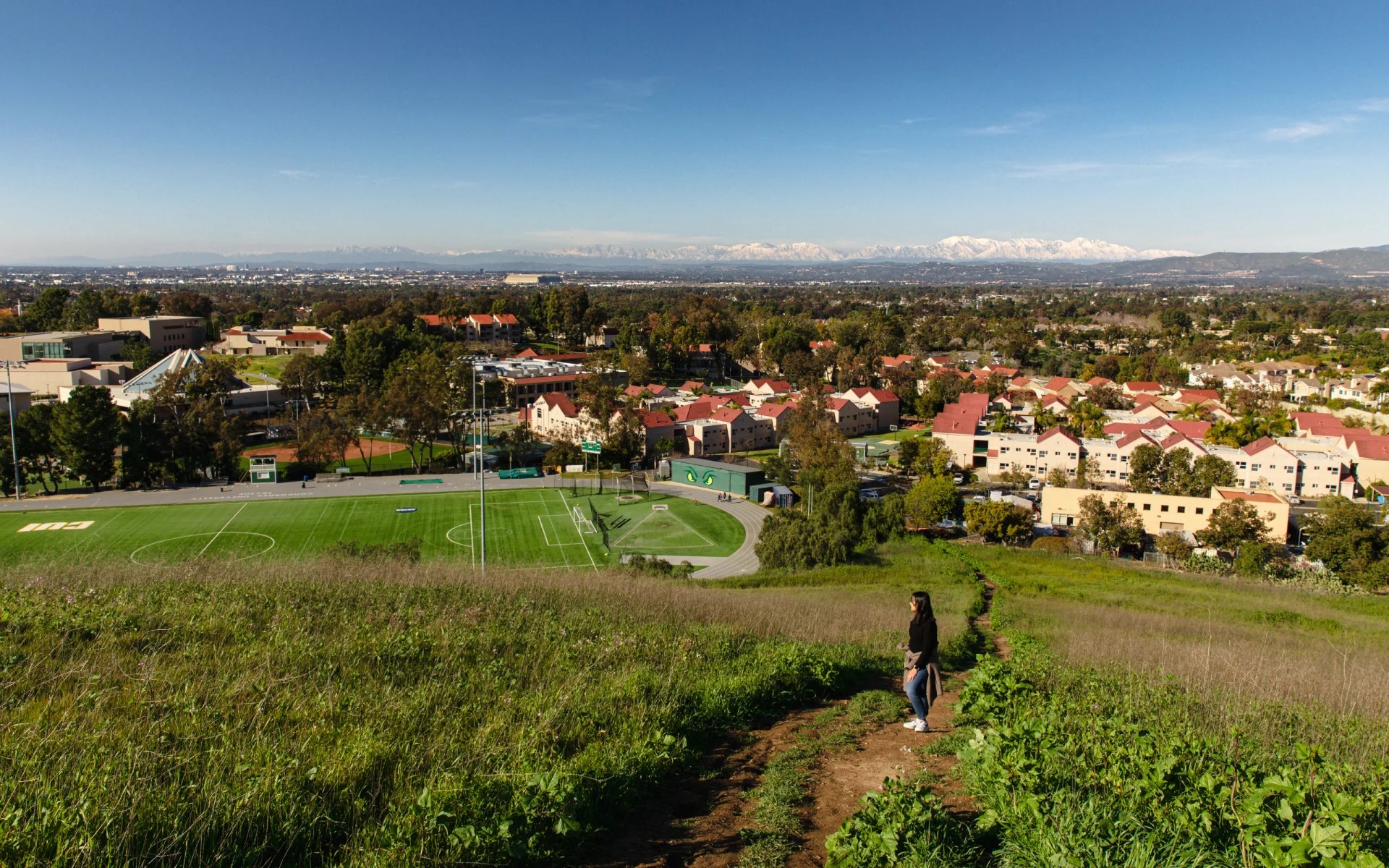 A person standing on a path in a grassy area with a football field and buildings in the background
