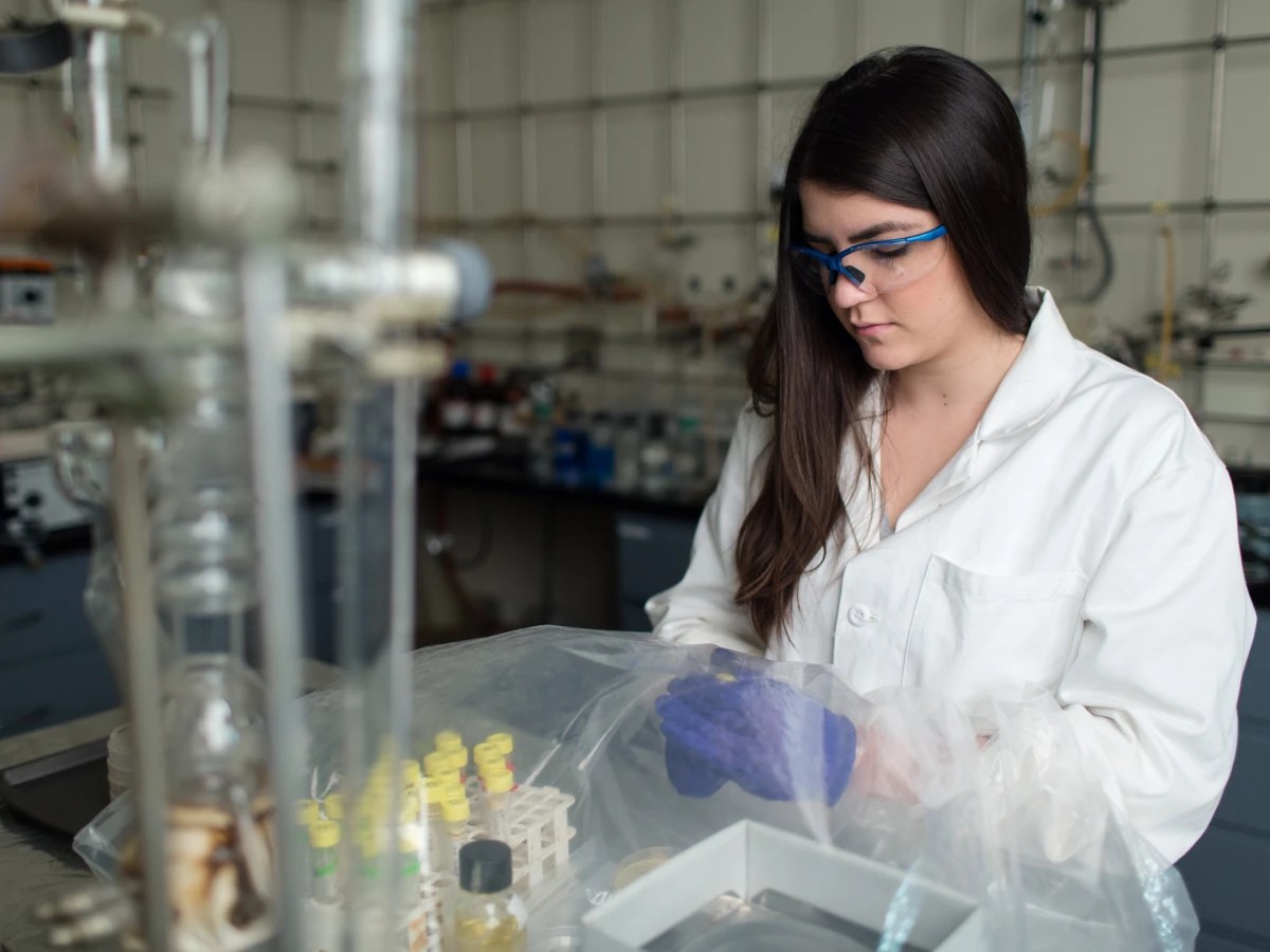 A person wearing a lab coat and safety glasses working with laboratory samples and glassware