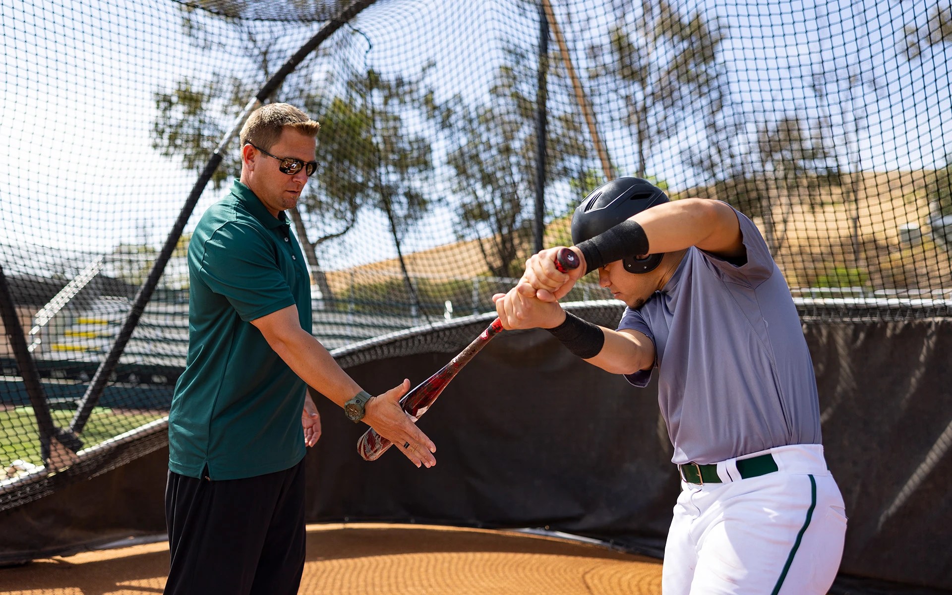 A coach guides a baseball player during batting practice, adjusting his swing inside a training cage
