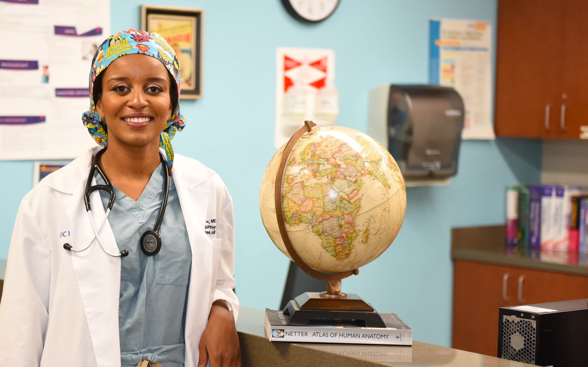 Person wearing scrubs and a lab coat standing by a globe in a medical setting