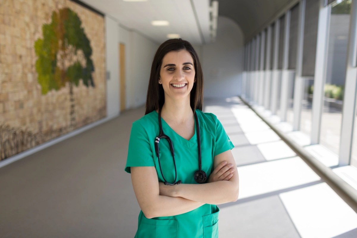 Person in green medical scrubs standing in a bright hallway with arms crossed and smiling