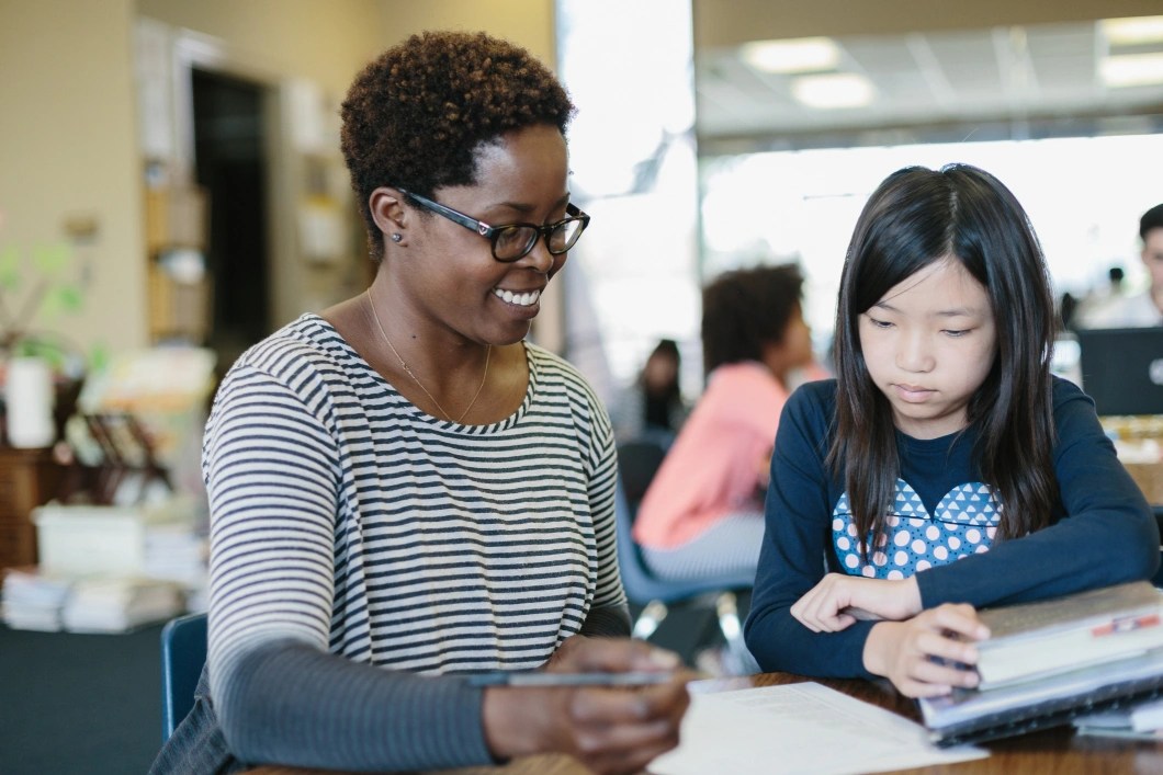 Teacher smiling while assisting a student with her schoolwork at a classroom table