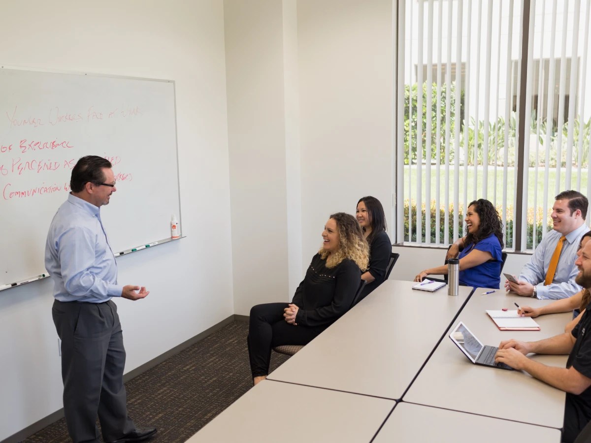 A person stands at the front of a classroom speaking to a group of people seated at tables, all smiling and engaged in conversation