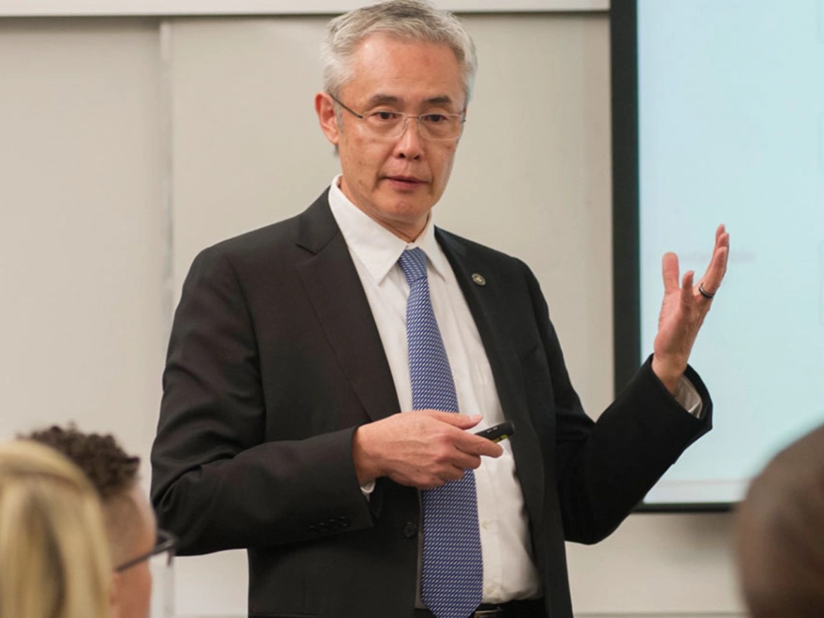 A person in a suit gives a presentation to a classroom, gesturing toward a projected slide while students listen attentively