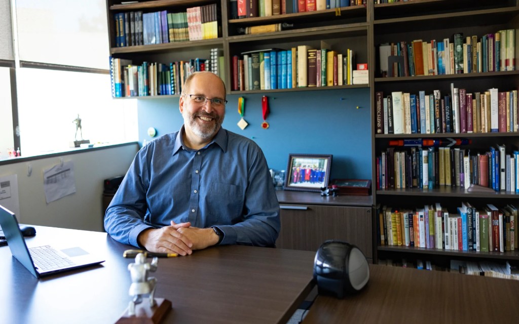 A man sitting at a desk with books on the wall behind him
