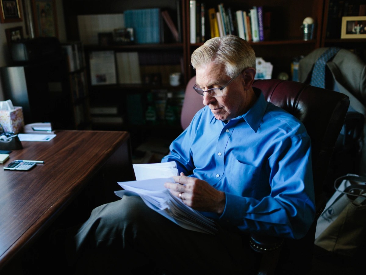 A person in a blue shirt sits at a desk reading papers in an office filled with books and documents at Concordia University Irvine