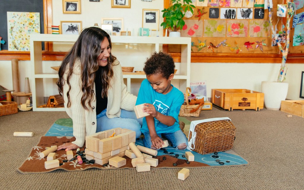 A school psychology student sits on the floor with a young child, smiling and playing with wooden blocks in a colorful classroom at Concordia University Irvine