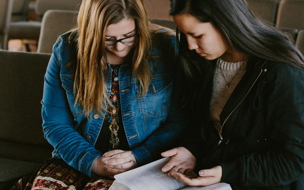 Two students sitting together and praying with an open Bible in a chapel setting