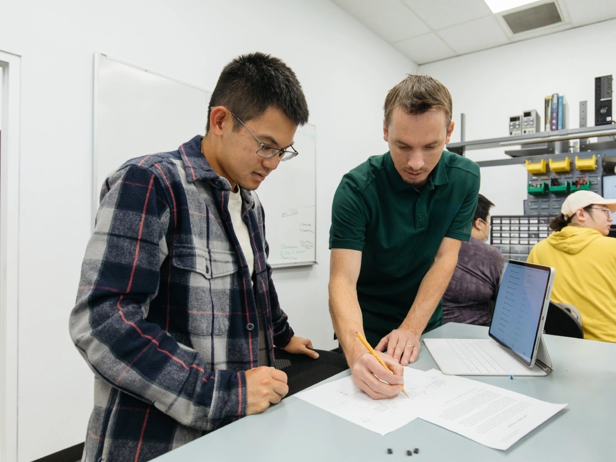 Two students working together on an electronic circuit design, reviewing schematics on paper beside a laptop in a lab setting