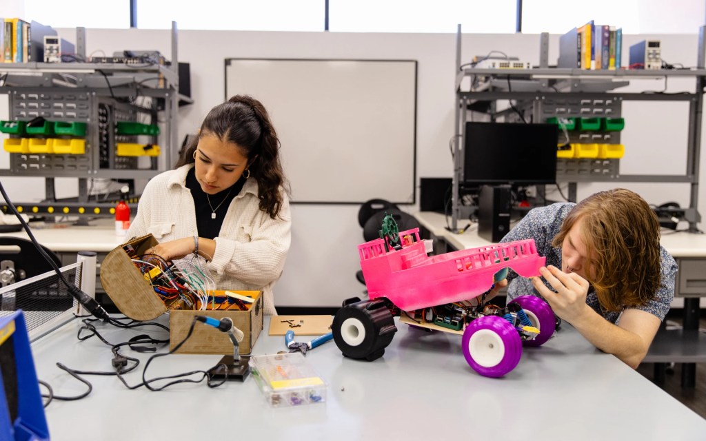 Two students working on an engineering project with electronic circuits and a pink robotic vehicle in a lab