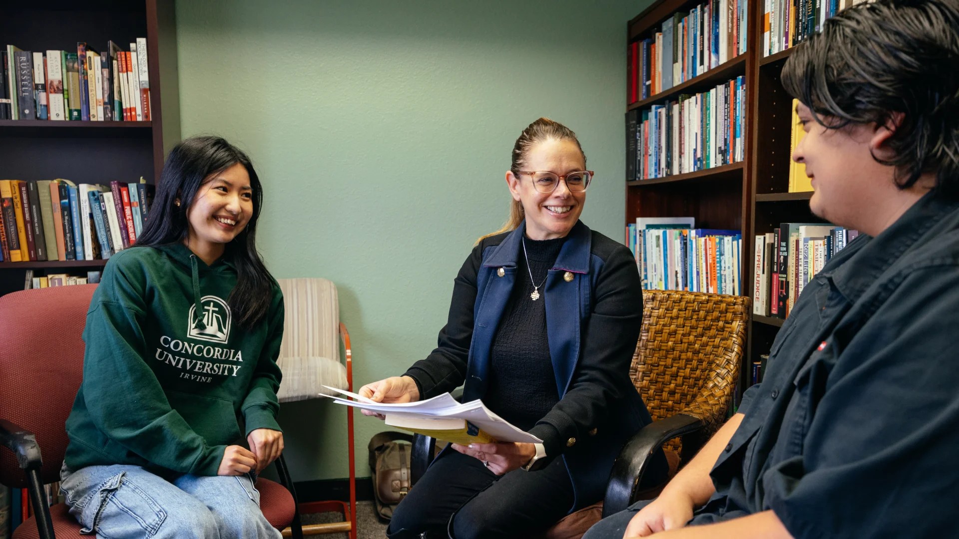 A professor meets with two students in an office lined with bookshelves, smiling and discussing papers