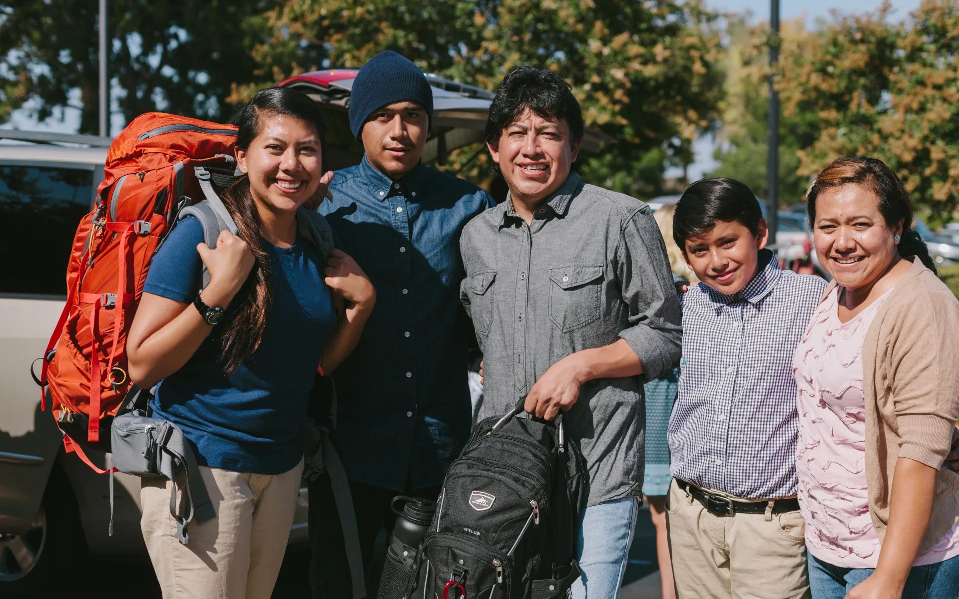 A family standing together outdoors, smiling as they help a student prepare for a trip with backpacks