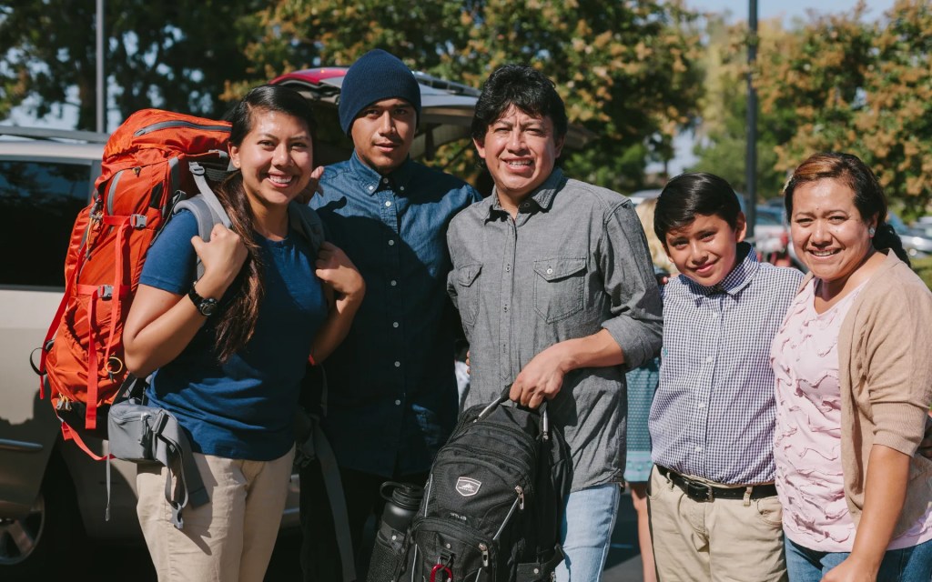 A family standing together outdoors, smiling as they help a student prepare for a trip with backpacks