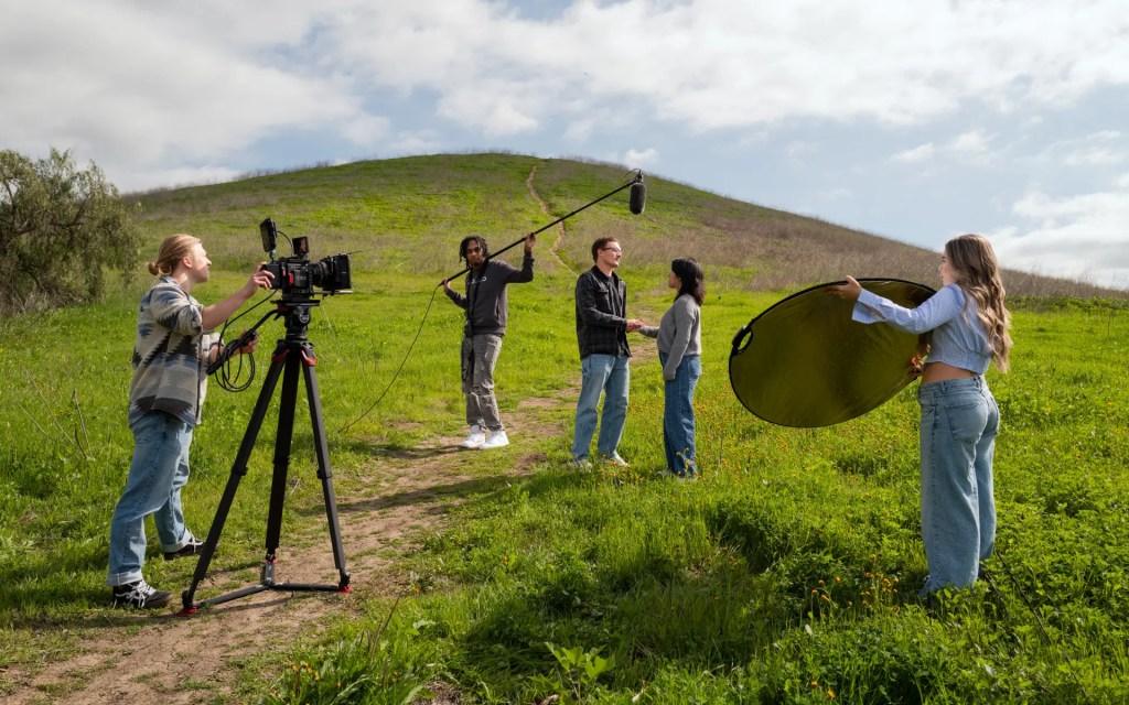 Group of film students recording an outdoor scene with a camera, boom mic, and reflector on a grassy hill