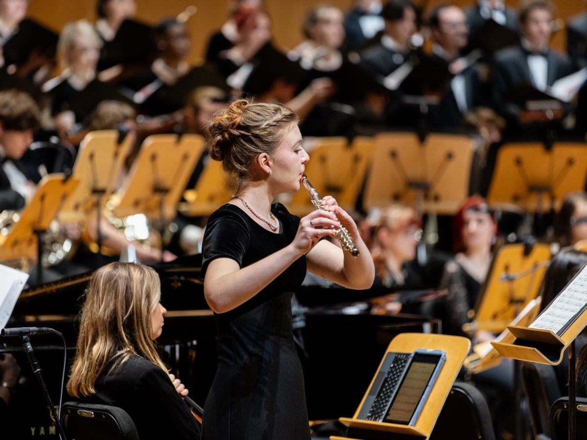 A musician playing the flute during a university orchestra concert, surrounded by other performers and music stands