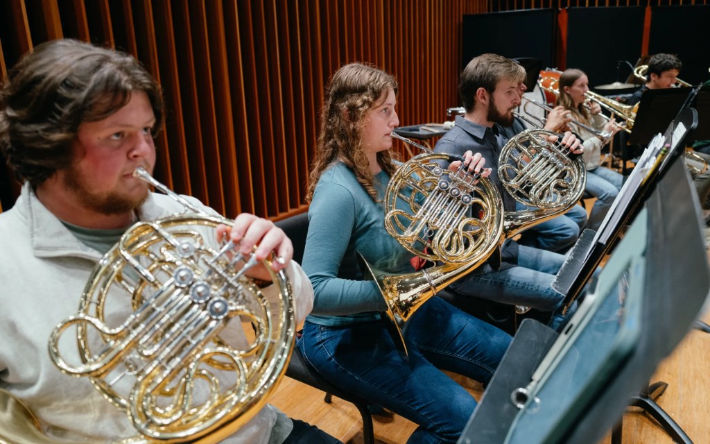 A group of university students playing French horns during an orchestra rehearsal in a music hall