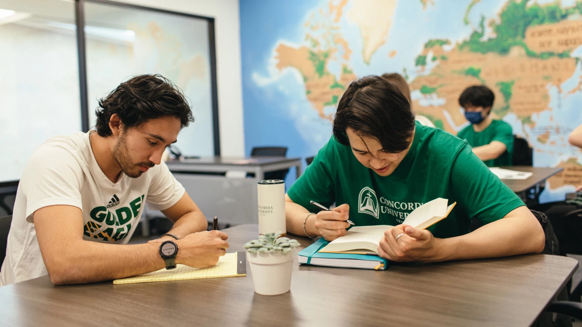 Two students studying together at a classroom table with books and notes, with a world map mural in the background