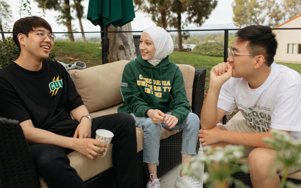 Three international students sitting outdoors on campus couches, smiling and chatting together