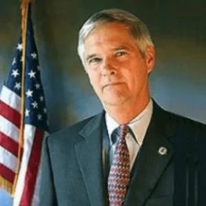 A man in a suit and tie standing in front of a flag