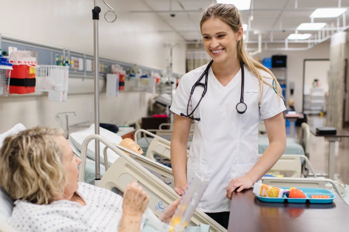 A person in scrubs standing beside a patient in a hospital bed, smiling and holding a medical device