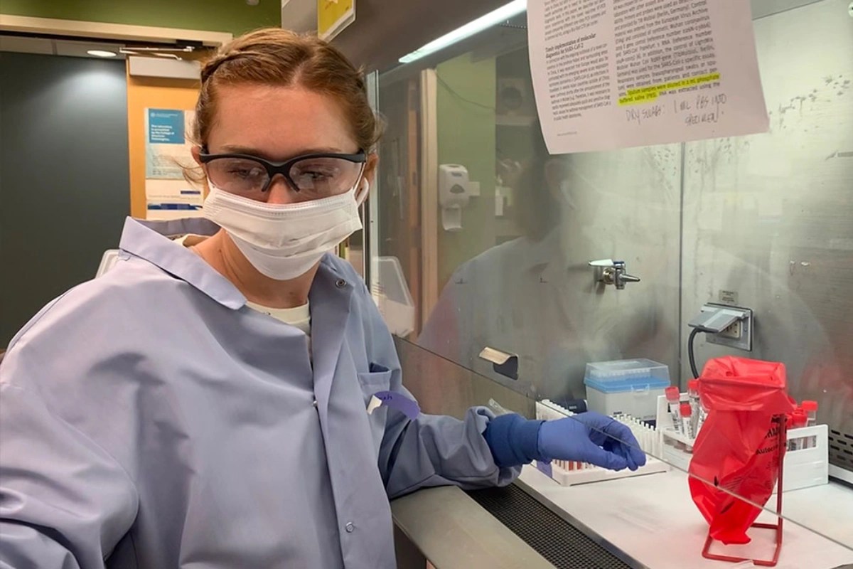 A person wearing a lab coat, mask, gloves, and goggles working under a fume hood with lab materials