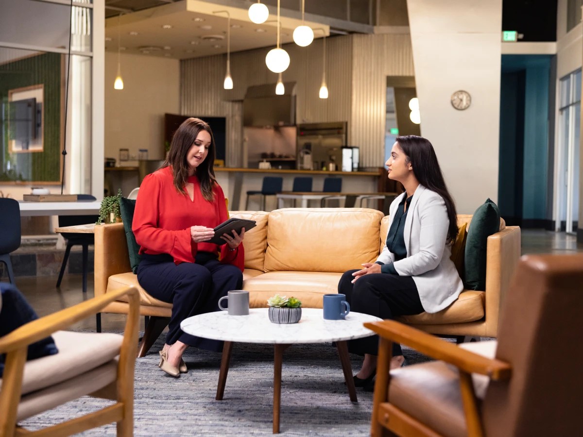 Two women in business attire having a professional meeting in a modern office lounge with coffee mugs on the table
