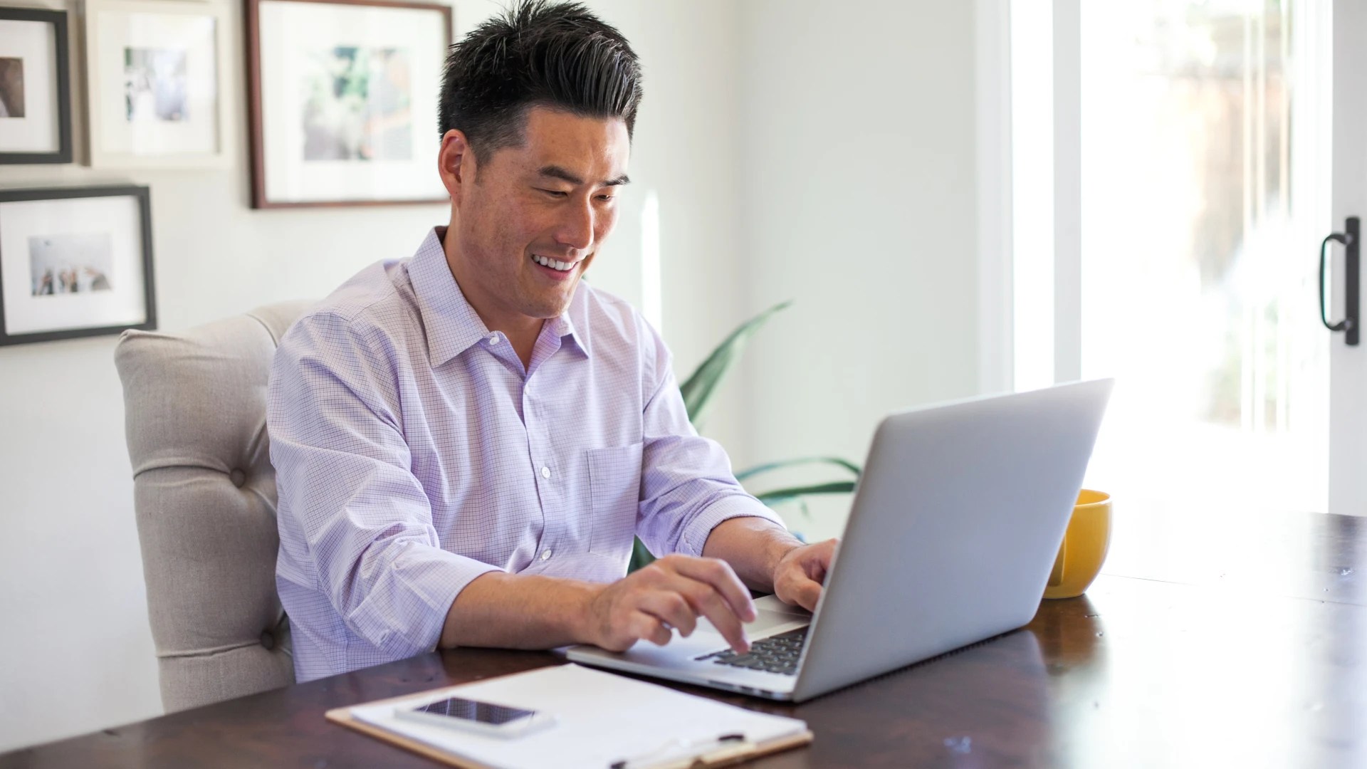Person sitting at a wooden desk using a laptop with a notebook, phone, and coffee mug nearby in a bright home office