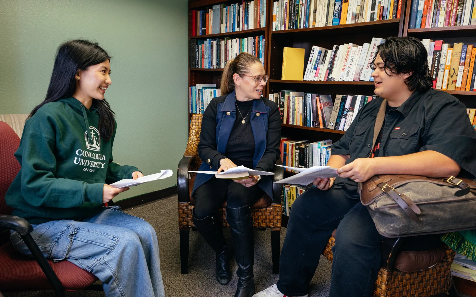 Professor meeting with two students in an office filled with bookshelves, discussing papers together