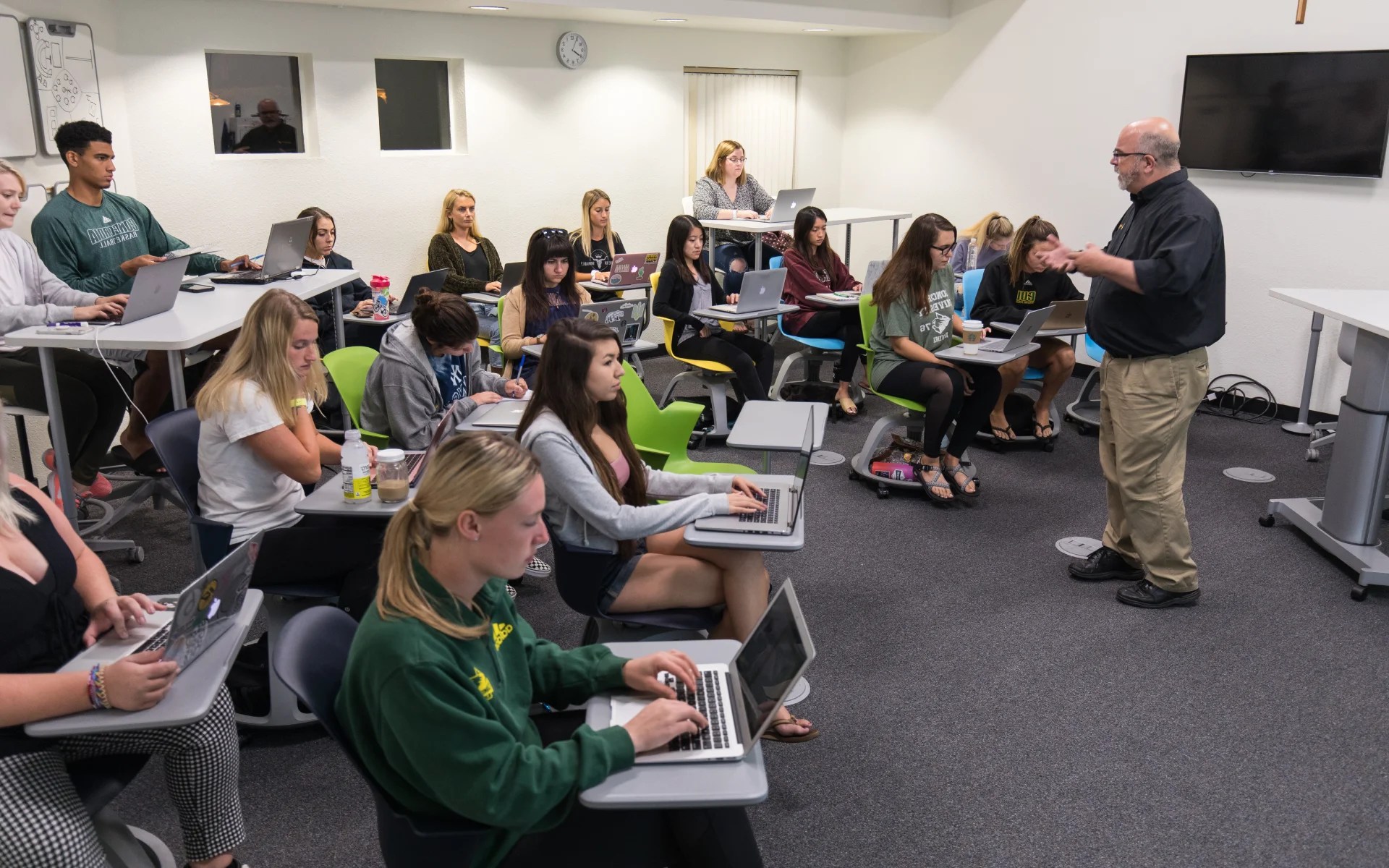 Professor teaching a class of college students using laptops in a modern, collaborative classroom setting