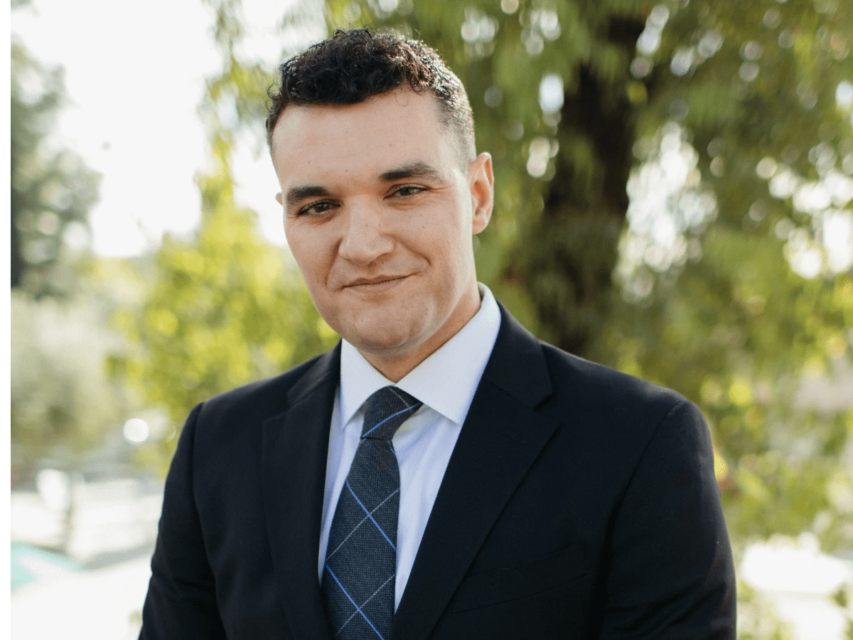 Person wearing a suit and tie standing outdoors with greenery in the background