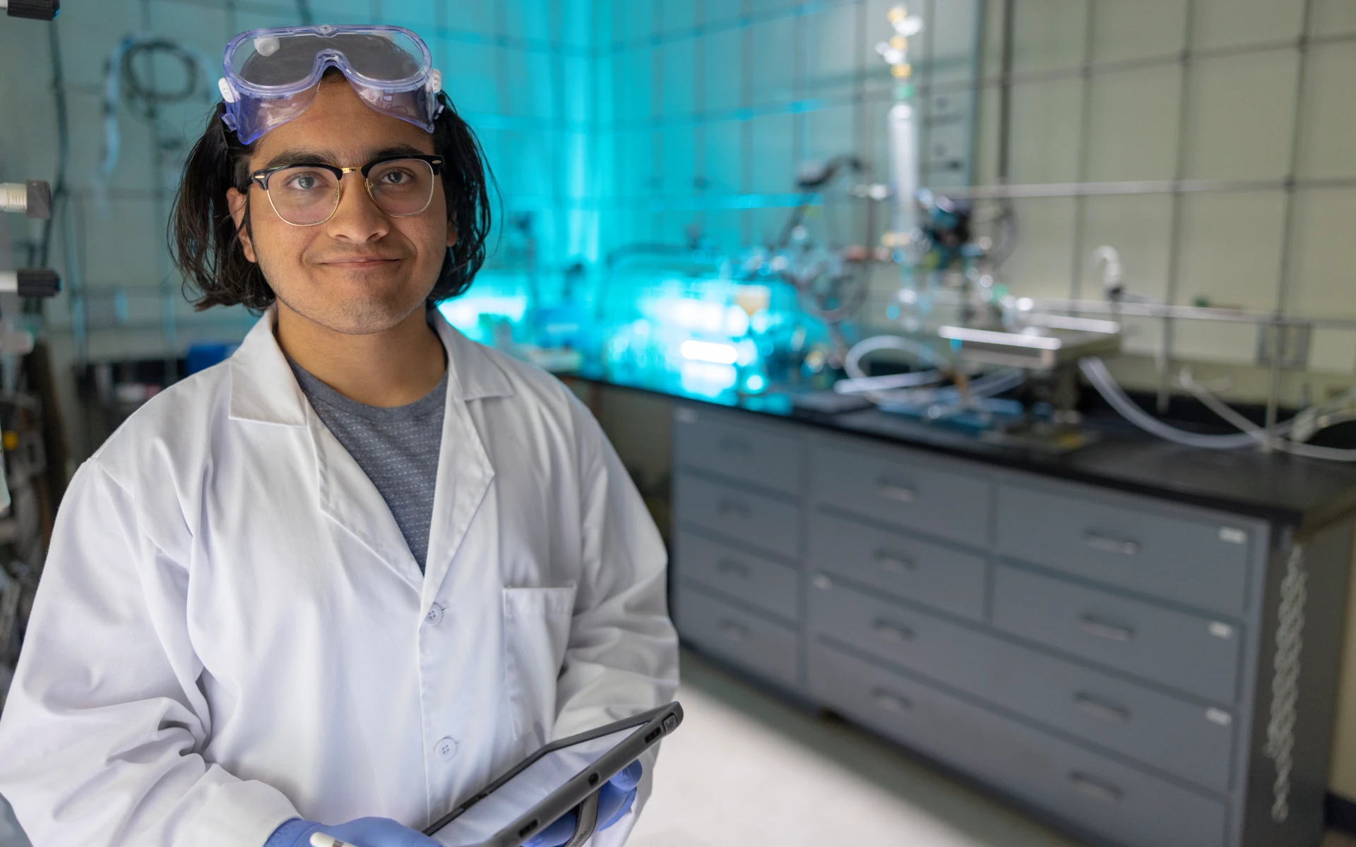 A person wearing a lab coat, gloves, and safety goggles holding a tablet in a laboratory setting