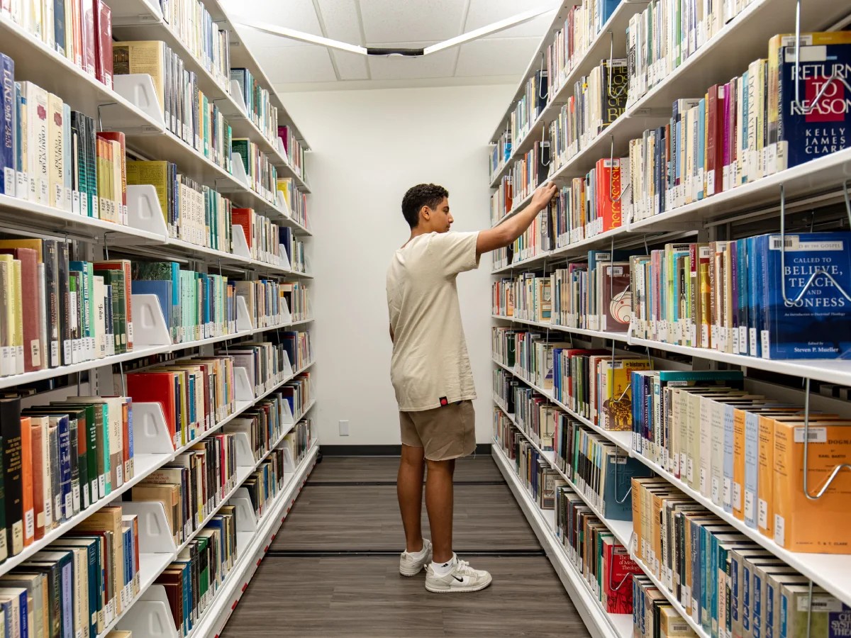Student standing between library shelves selecting a book from a row of colorful volumes