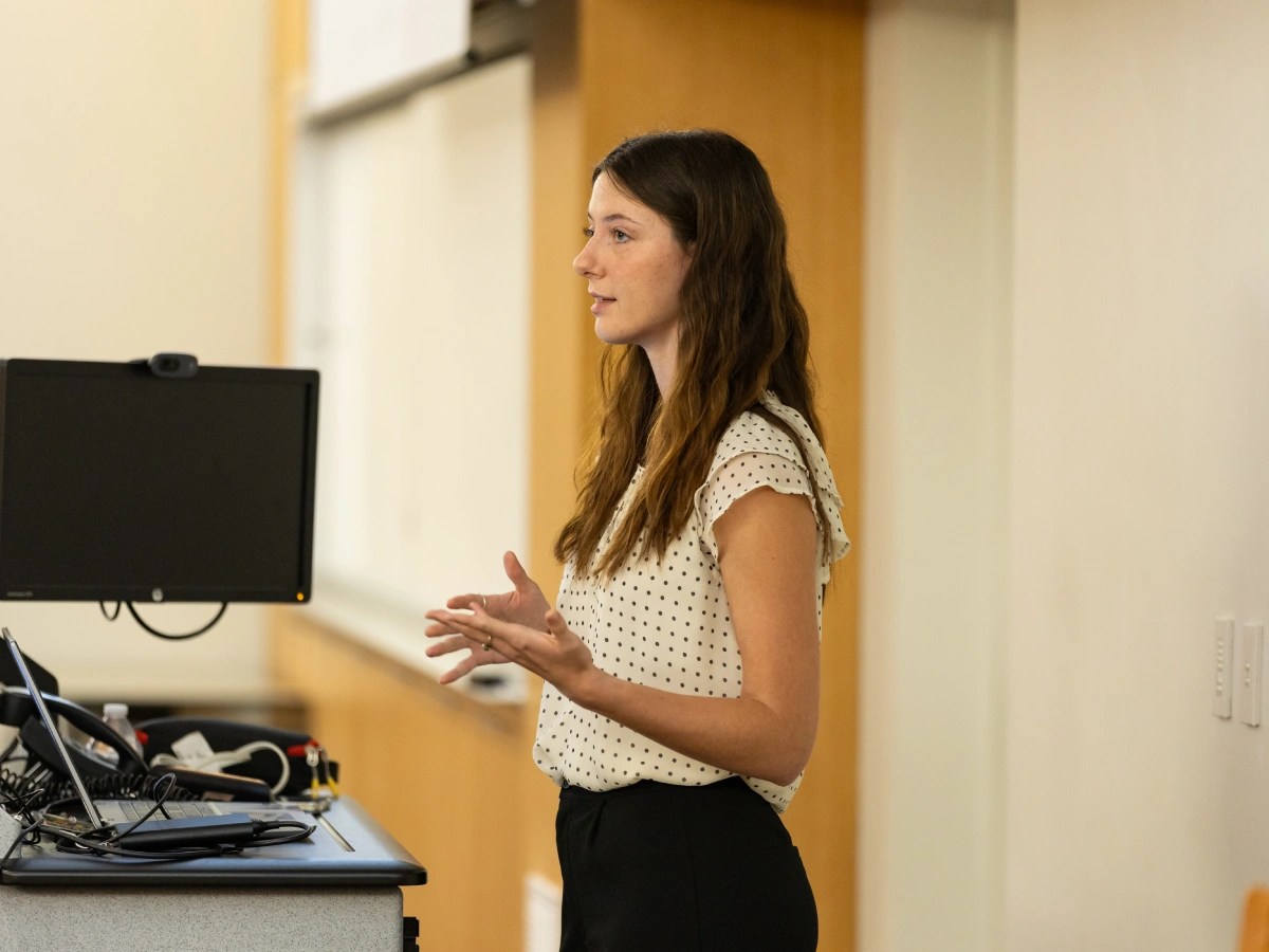 Student giving a presentation in a classroom, standing near a computer and speaking to the audience