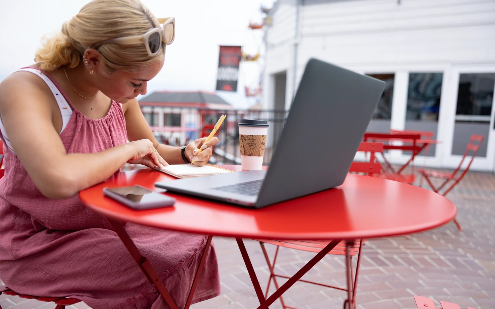A person writing in a notebook while studying outdoors at a red café table with a laptop, coffee cup, and phone nearby