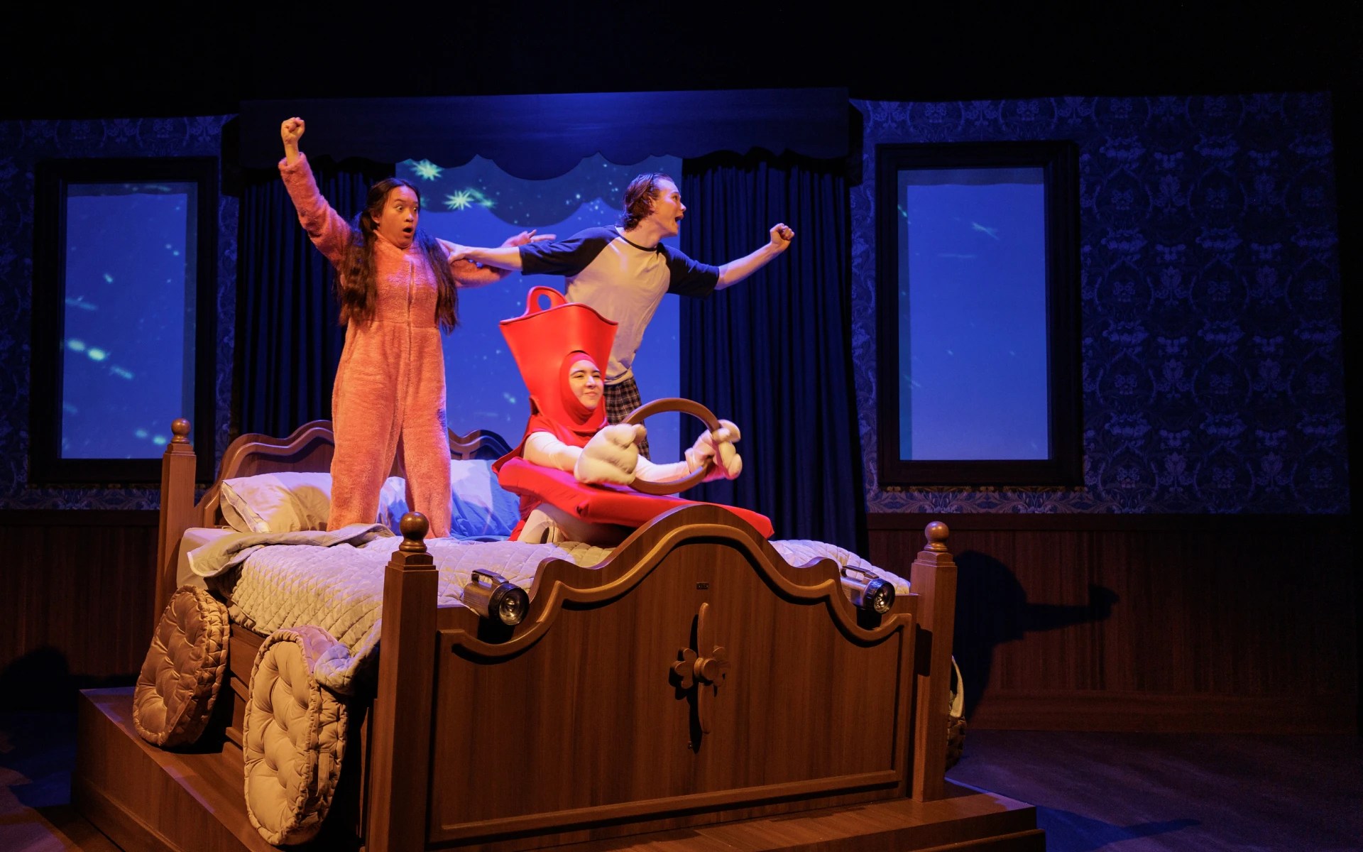 Students performing in a colorful stage play with a large bed prop and creative costumes