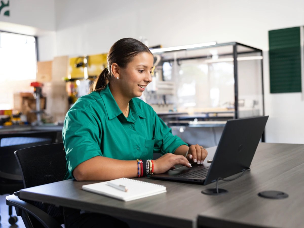 Person sitting at a table using a laptop in a modern lab or classroom setting with notebooks nearby