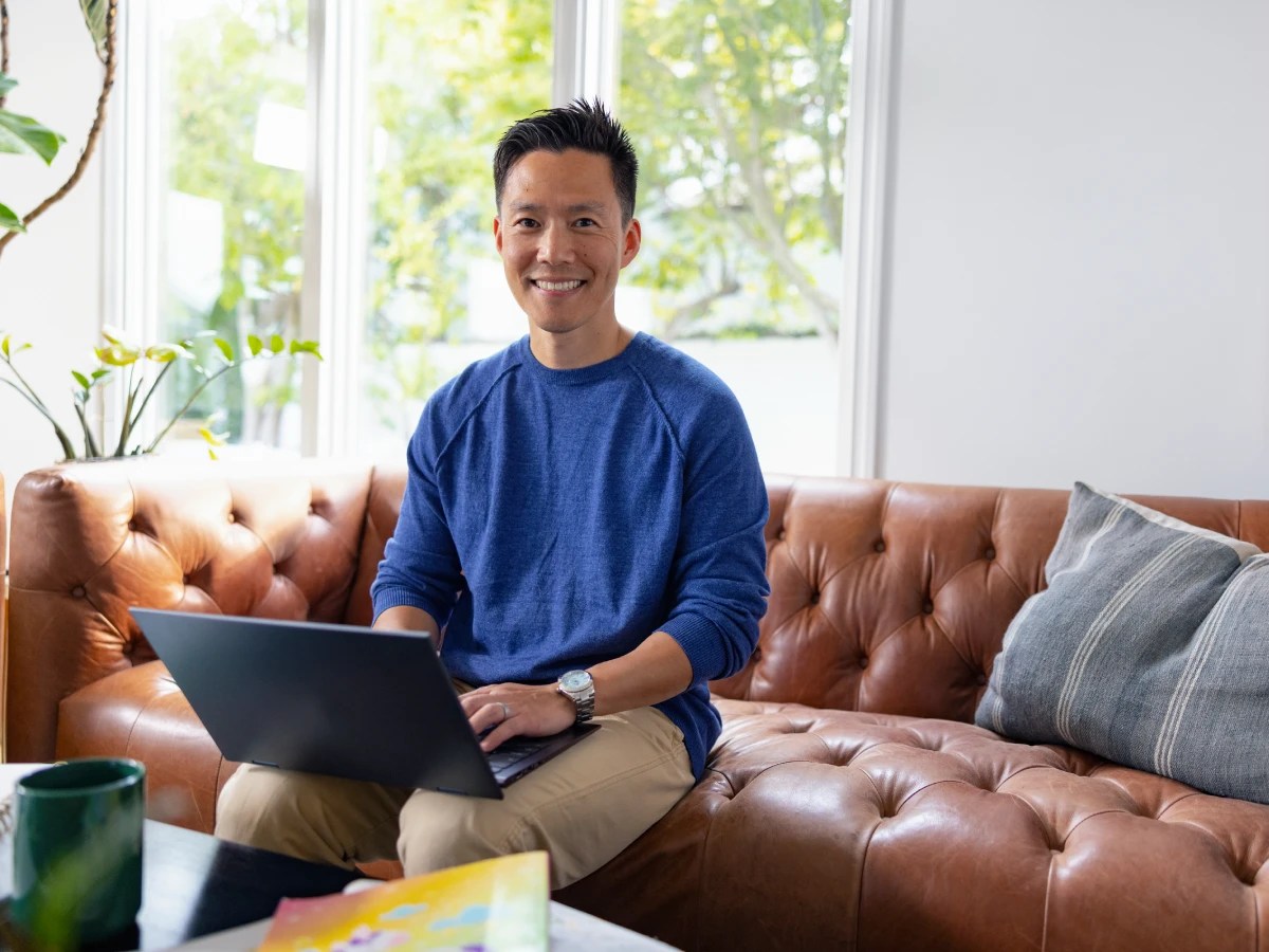 A person smiling while working on a laptop from a couch in a bright living room with natural light