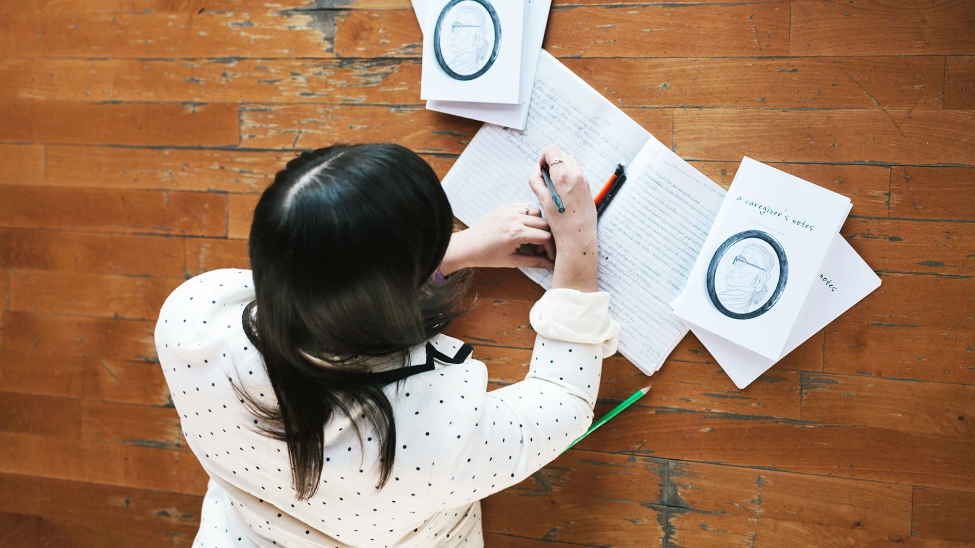Student writing in a notebook on a wooden floor, surrounded by open journals and papers