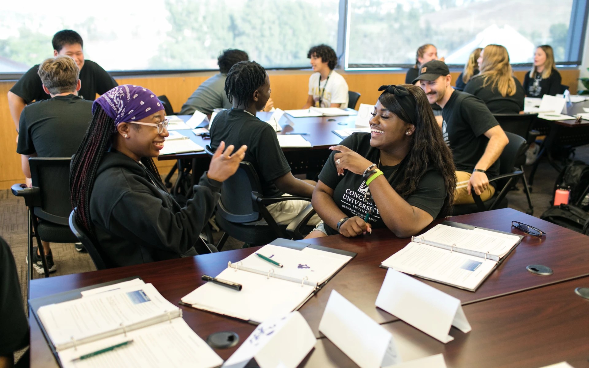 A group of people sitting around tables with binders, laughing and talking during a classroom session
