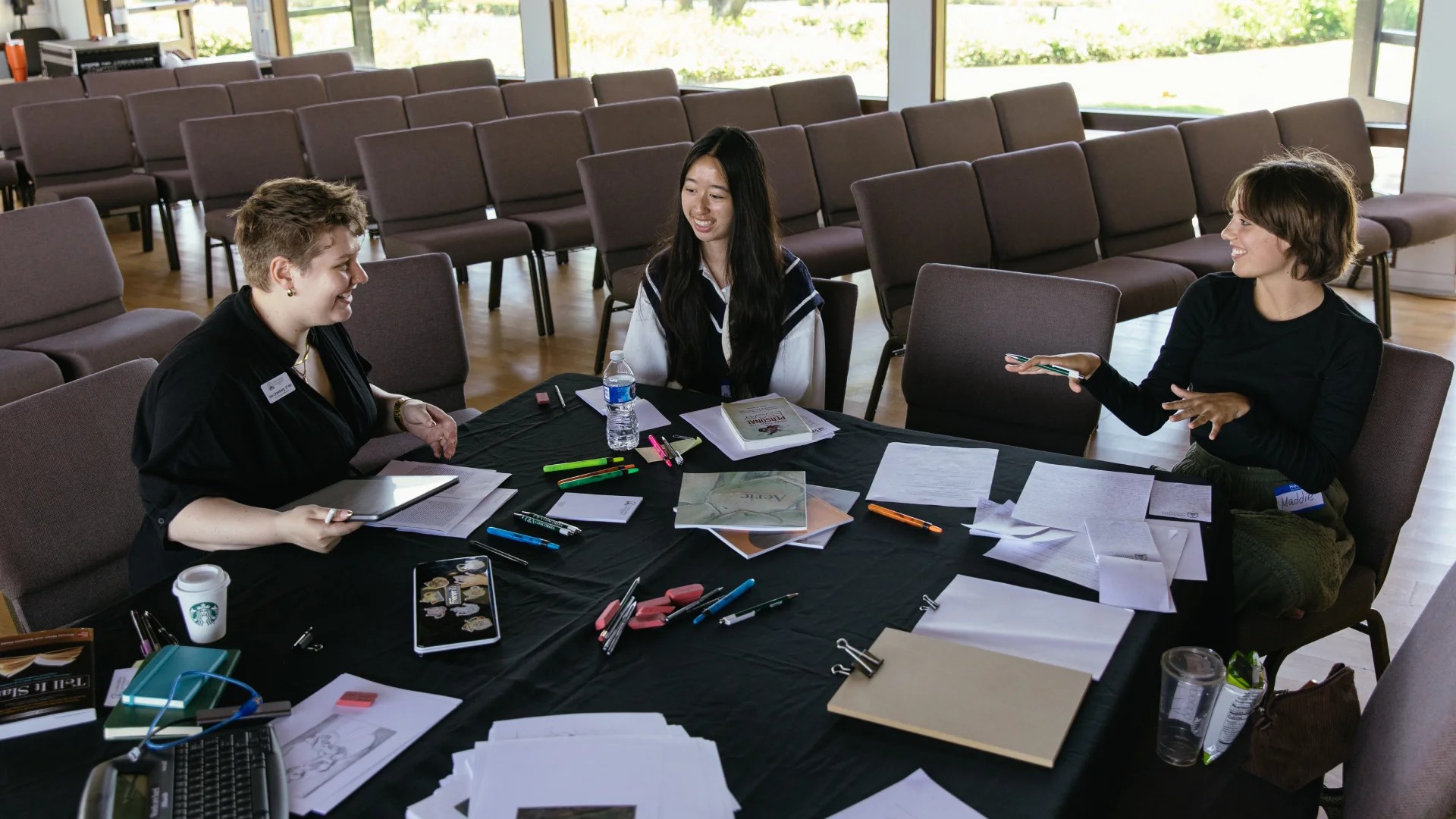 Three students sitting at a table covered with notebooks, pens, and papers, collaborating during a creative arts workshop