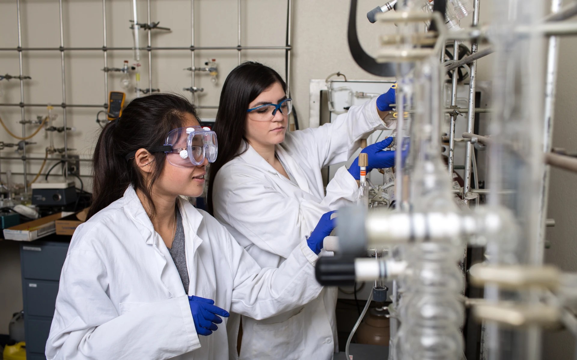 Two people wearing lab coats, gloves, and safety goggles conducting an experiment with glass lab equipment