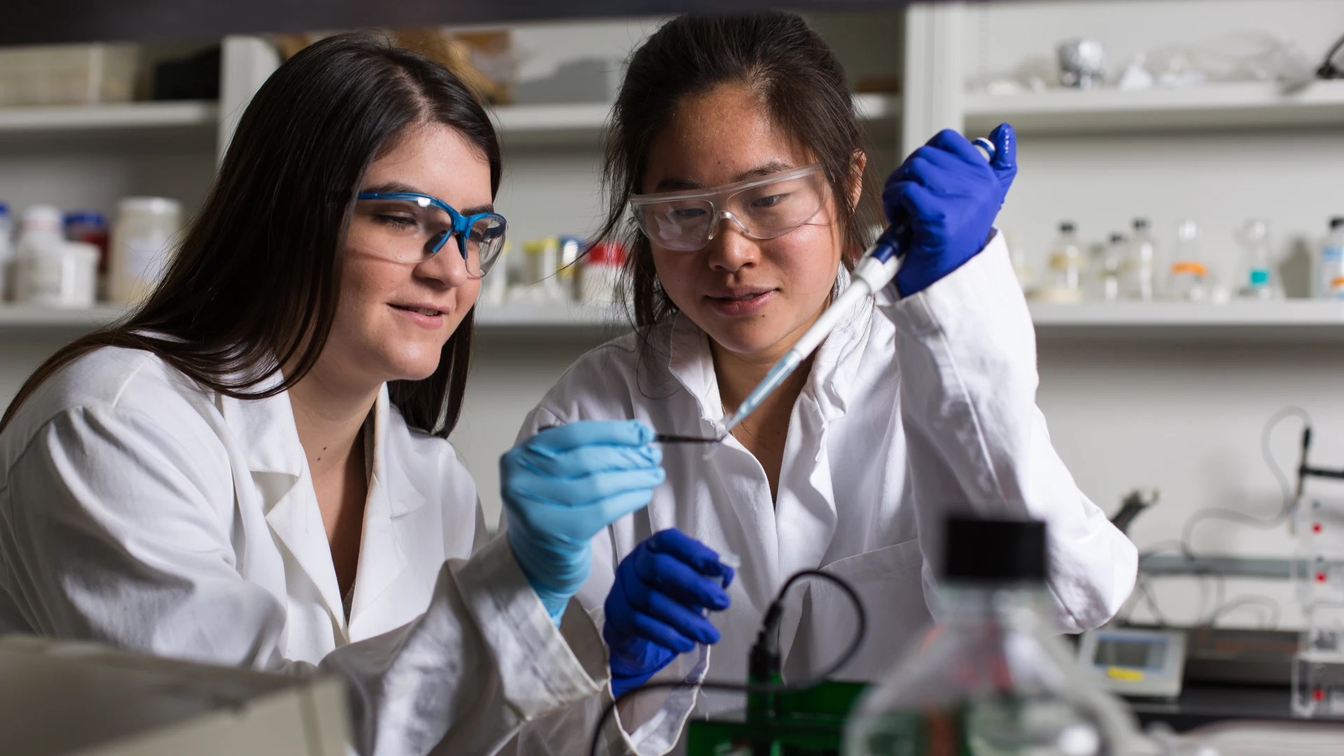 Two people wearing lab coats and safety goggles working together with lab equipment and pipettes