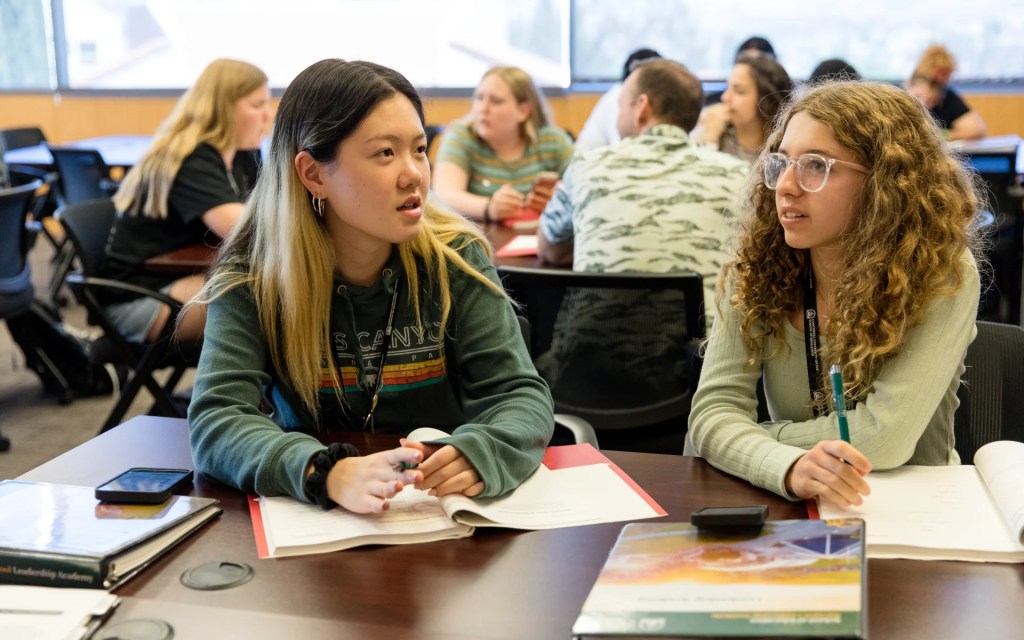 Two students sit at a classroom table, talking and taking notes during a group discussion