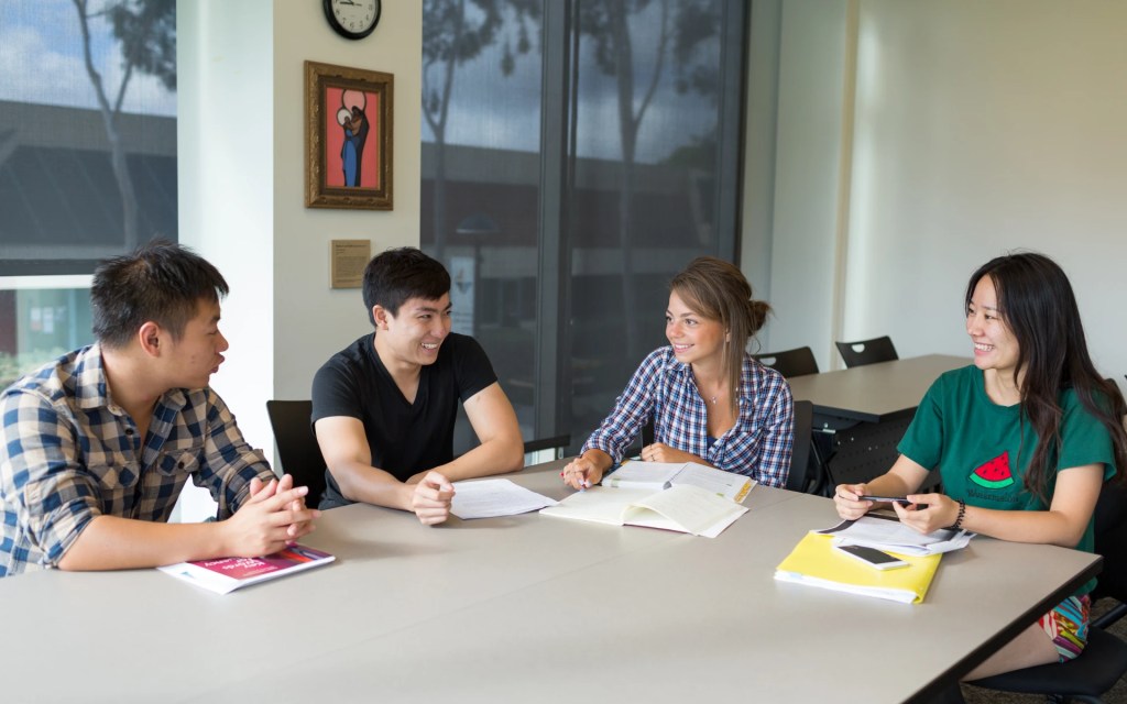 Four students sitting around a table in a classroom working together on an assignment and smiling