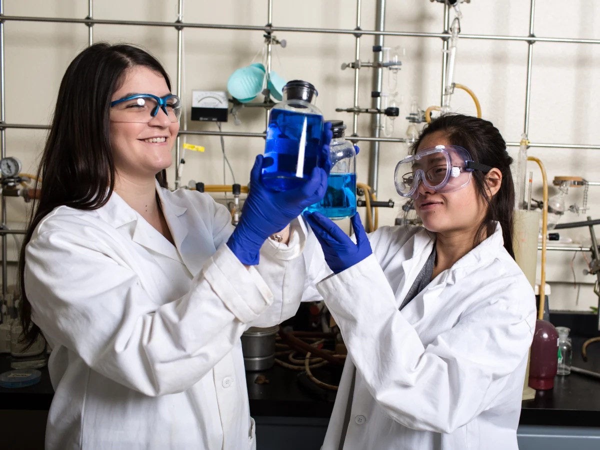 Two people wearing lab coats, gloves, and goggles working together to mix a blue chemical solution in a laboratory