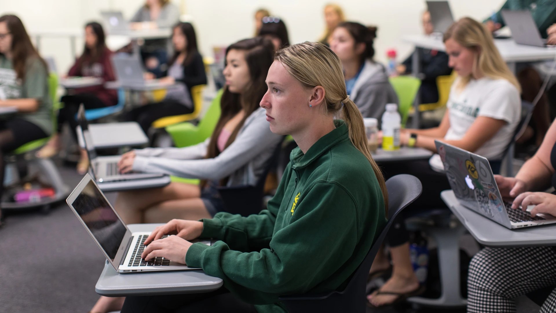 Students attentively listening in a classroom while working on laptops