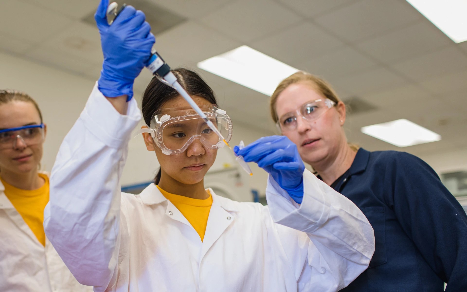 Three people wearing lab coats, gloves, and goggles working with a pipette in a laboratory setting
