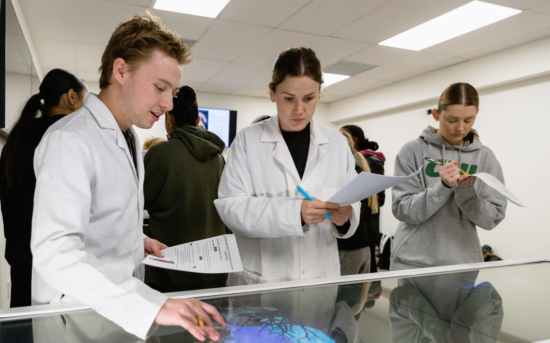 People wearing lab coats working with an interactive anatomy table and taking notes in a classroom
